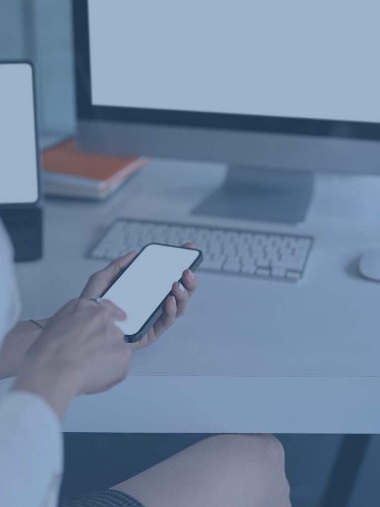 Person holding cell phone with other computer screens in background.